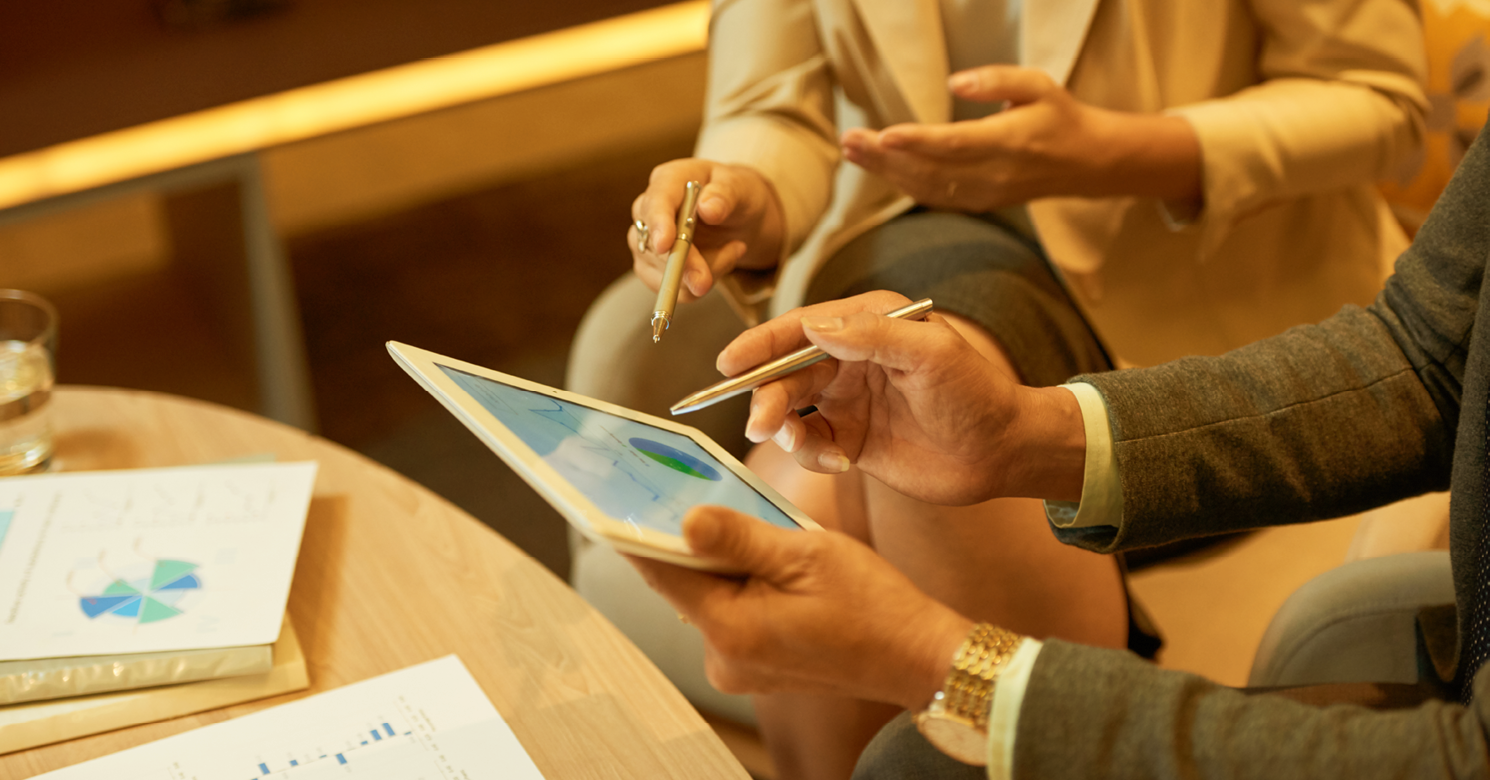Pessoas seguram canetas de tablet e o próprio aparelho, observando gráficos. Imagem ilustra matéria "Taxa máxima de juros do consignado sobe para 1,80% ao mês".